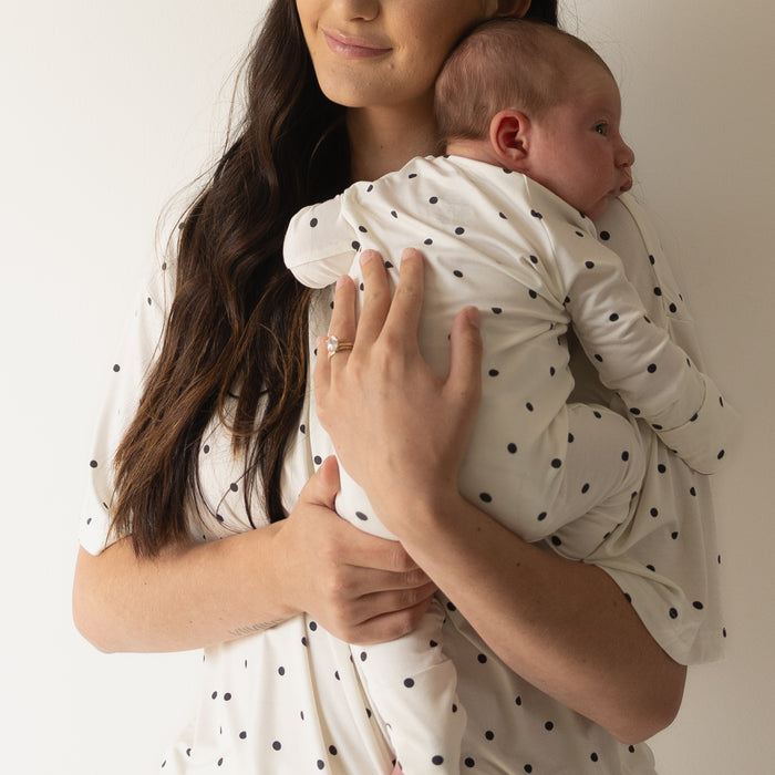 A woman with long brown hair holds a baby against her chest, both dressed in forever french baby's Bamboo Zip Pajamas | Annie—matching white outfits with black polka dots made from breathable, comfy sleepwear. The baby rests sideways on her shoulder.