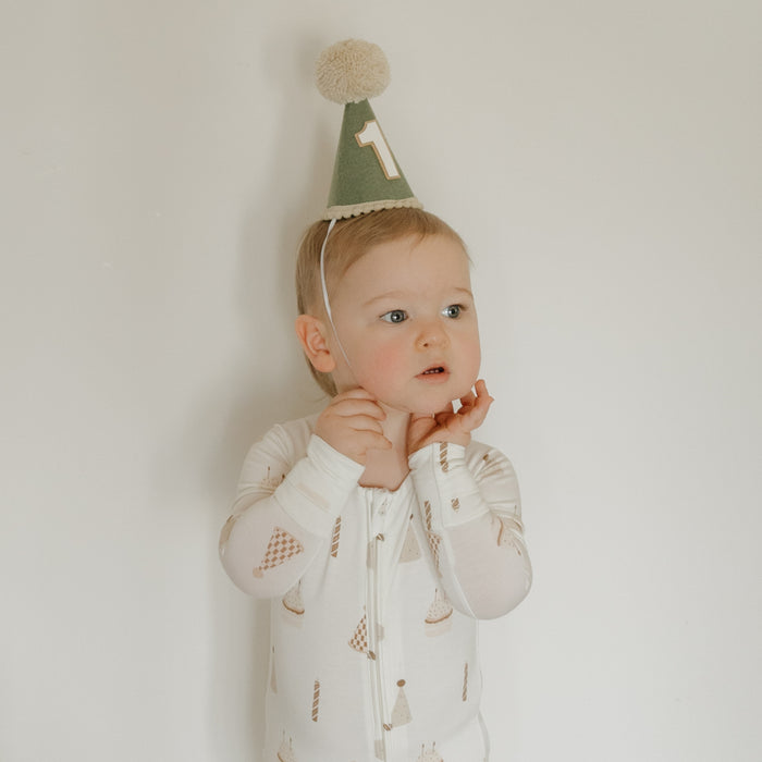 A toddler wearing forever french baby's Bamboo Zip Pajamas | Birthday Boy—a hypo-allergenic, white onesie adorned with ice cream cone prints—stands against a plain white wall, looking off to the side with hands near their face and sporting a green party hat with a “1” on it.