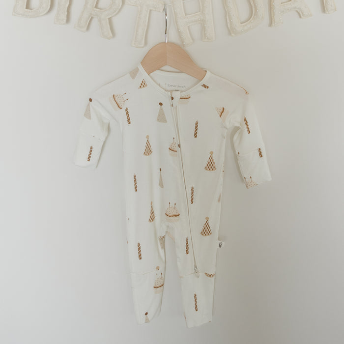 A pair of white "Bamboo Zip Pajamas | Birthday Boy" by forever french baby, made from hypoallergenic bamboo fabric and featuring birthday-themed prints like cake and candles, hangs on a wall beneath a cream-colored "BIRTHDAY" banner.