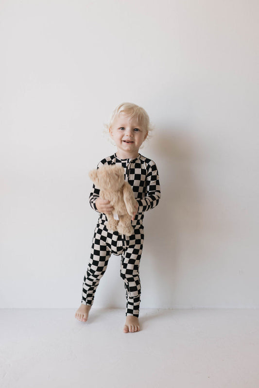 A small child with curly blond hair is standing against a plain white background, wearing forever french baby's Bamboo Zip Pajamas in the Black Checkerboard pattern featuring rollover cuffs. The child is barefoot, holding a light brown teddy bear, and smiling at the camera.