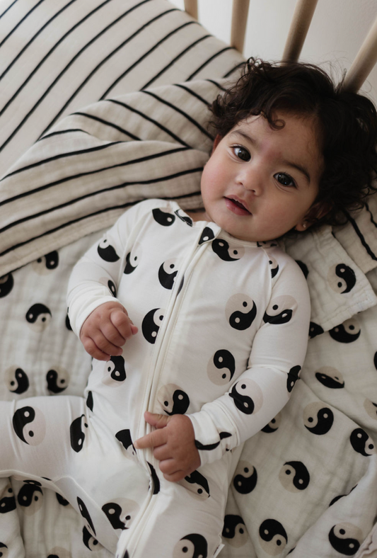 A baby with curly hair is lying on a bed, dressed in "Bamboo Zip Pajamas | Black & White Yin Yang" by forever french baby. The bed’s bedding also features a similar yin-yang design along with black and white stripes, creating a serene setting. The baby is looking towards the camera.