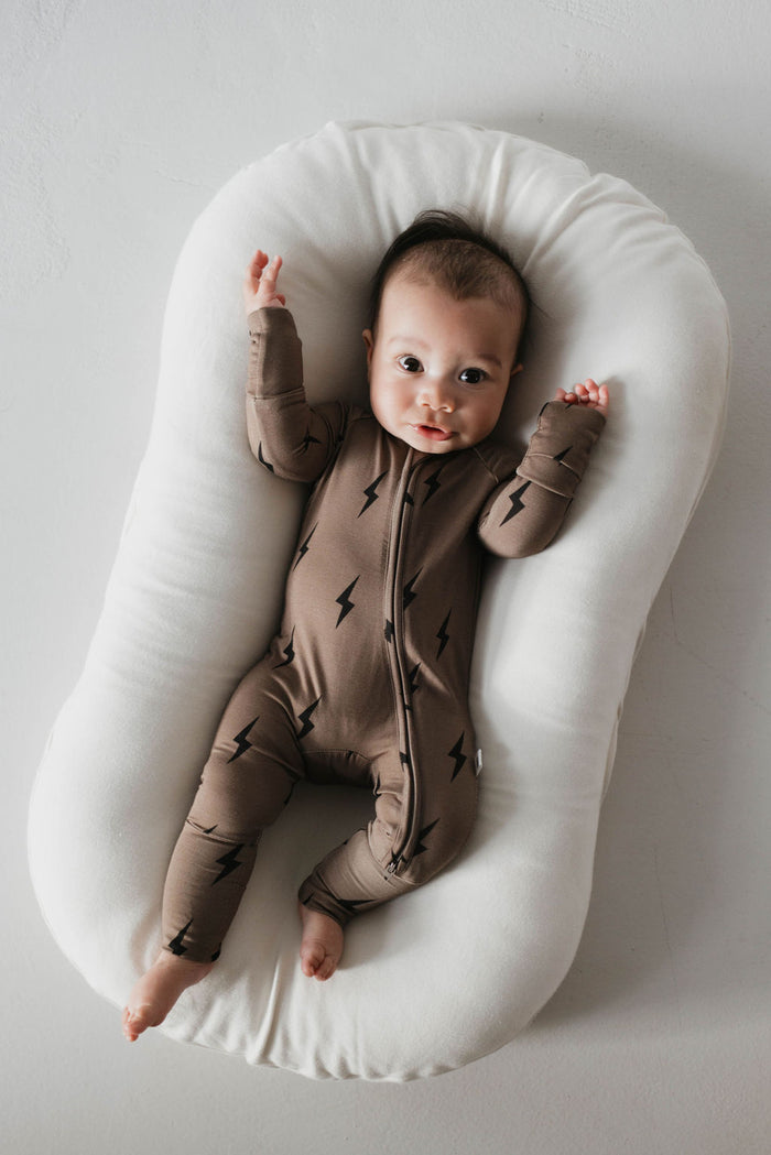 A baby lies on a white, cushioned surface wearing the Bamboo Zip Pajamas adorned with a brown and black lightning bolt pattern from Forever French Baby. The baby's arms are stretched above their head, and they are looking towards the camera with an alert expression. This hypo-allergenic outfit ensures both style and comfort.
