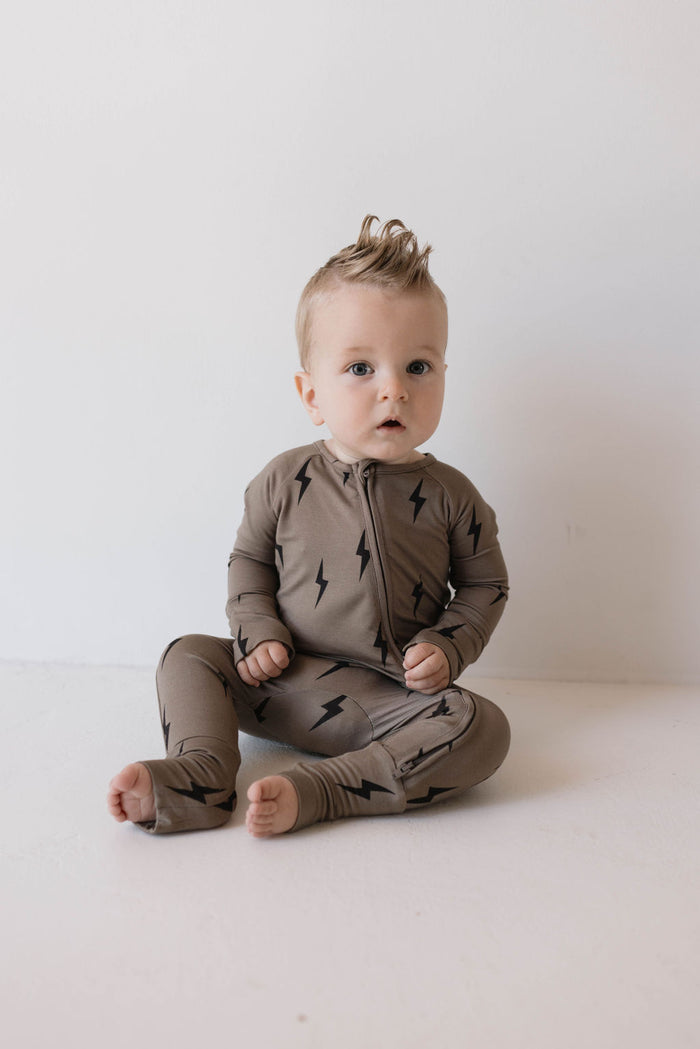 A baby with short, light-colored hair sits on the floor against a white background. The baby is wearing Bamboo Zip Pajamas in Brown & Black Lightning Bolt from forever french baby, made from soft premium bamboo that is hypo-allergenic. The baby looks forward with a relaxed expression.