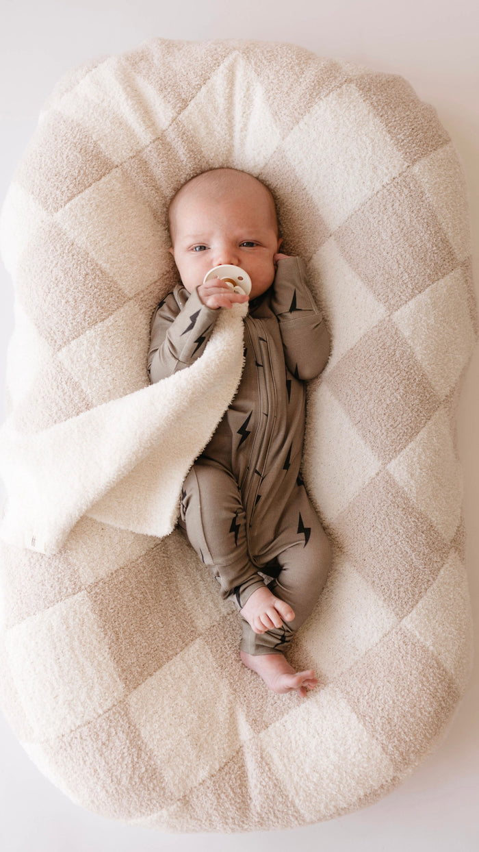 A baby dressed in forever french baby's Bamboo Zip Pajamas in a brown and black lightning bolt pattern lies on a plush, checkered cushion. The baby's left hand rests on their face while the right hand holds a pacifier. A white, hypo-allergenic blanket partially covers the baby.