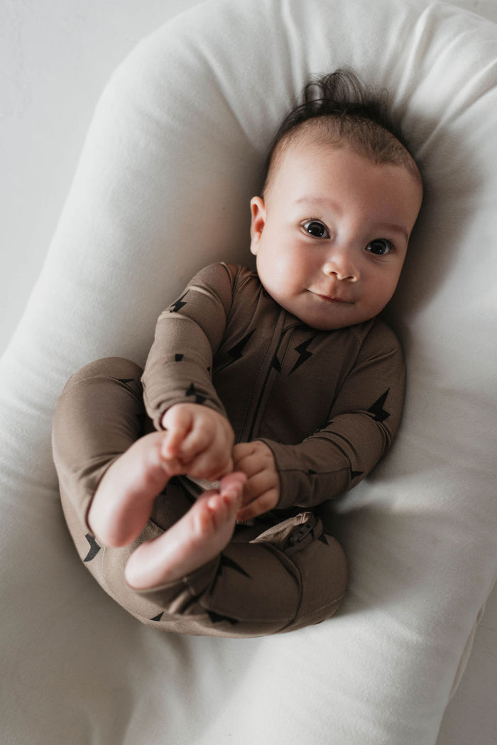 A baby dressed in Forever French Baby's Brown & Black Lightning Bolt Bamboo Zip Pajamas lies on their back on a white cushion. With dark hair and wide eyes looking up with a slight smile, the baby holds their feet with their hands. These hypoallergenic pajamas ensure gentle comfort on the baby's skin.