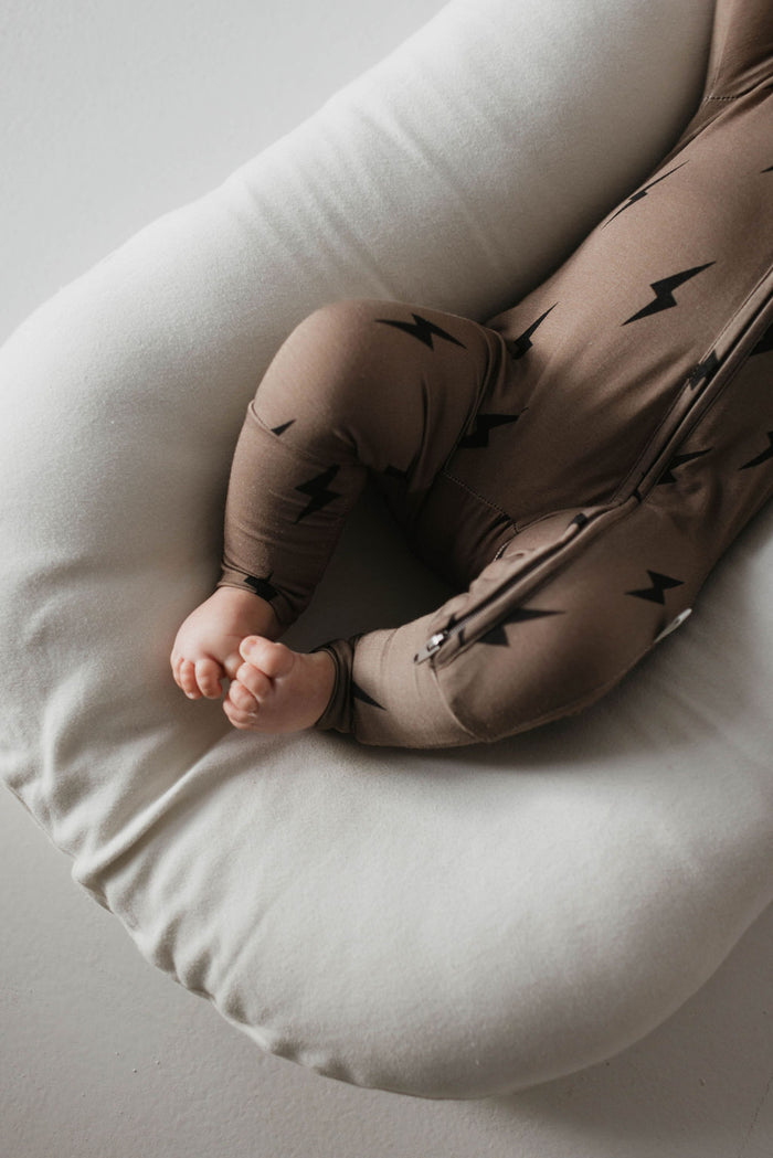 A baby lies on a soft, white cushion, dressed in the Bamboo Zip Pajamas | Brown & Black Lightning Bolt from forever french baby. The image highlights the baby's legs and tiny feet, partially visible against a minimal, light-colored background. The hypo-allergenic outfit promises comfort with its soft premium bamboo fabric.