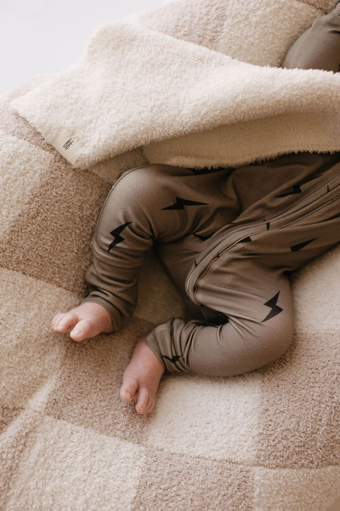 A small baby lies down, partially covered with a cozy beige and cream checkered blanket. Wearing forever french baby's Bamboo Zip Pajamas featuring brown leggings with black lightning bolt designs, only the baby’s legs and feet are visible.