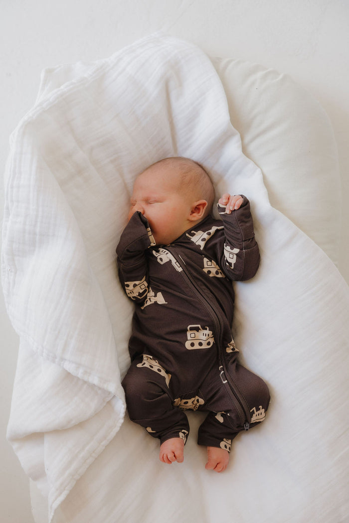 A newborn baby sleeps on a white blanket wearing forever french baby Bamboo Zip Pajamas | Bulldozer—a dark onesie with light vehicle patterns—and rests one hand near their face.