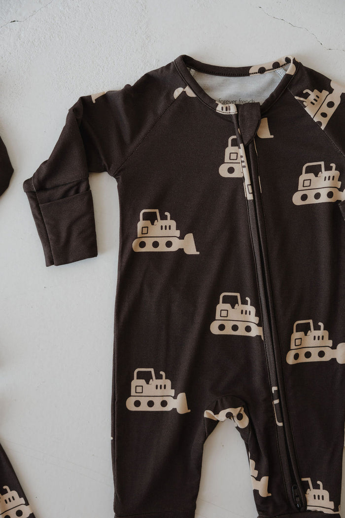 The forever french baby Bamboo Zip Pajamas | Bulldozer feature a dark brown, long sleeve onesie with a beige bulldozer pattern and zipper, made from breathable fabric and shown laid flat on a light surface.