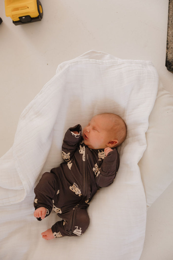 A newborn in forever french baby’s Bamboo Zip Pajamas | Bulldozer sleeps peacefully on a soft white blanket, with a touch of yellow visible nearby.