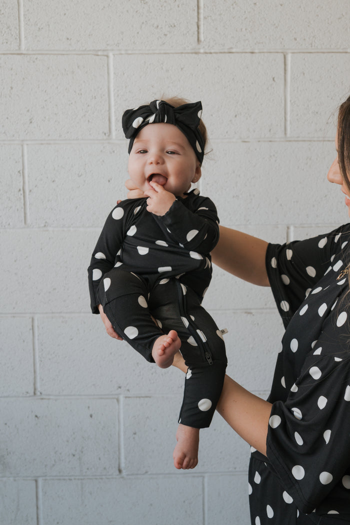 A baby dressed in forever french baby’s Bamboo Zip Pajamas in the B&W Polka Dot pattern, complete with a matching headband, is held by an adult whose arm and partial body are also clad in coordinating polka dot attire. Both wear the hypo-allergenic outfits against a white brick wall backdrop.
