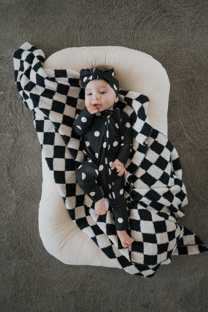 A baby wearing forever french baby’s Bamboo Zip Pajamas | B&W Polka Dot and a matching headband rests on a white cushion, wrapped in a black and white checkered blanket on a gray floor.