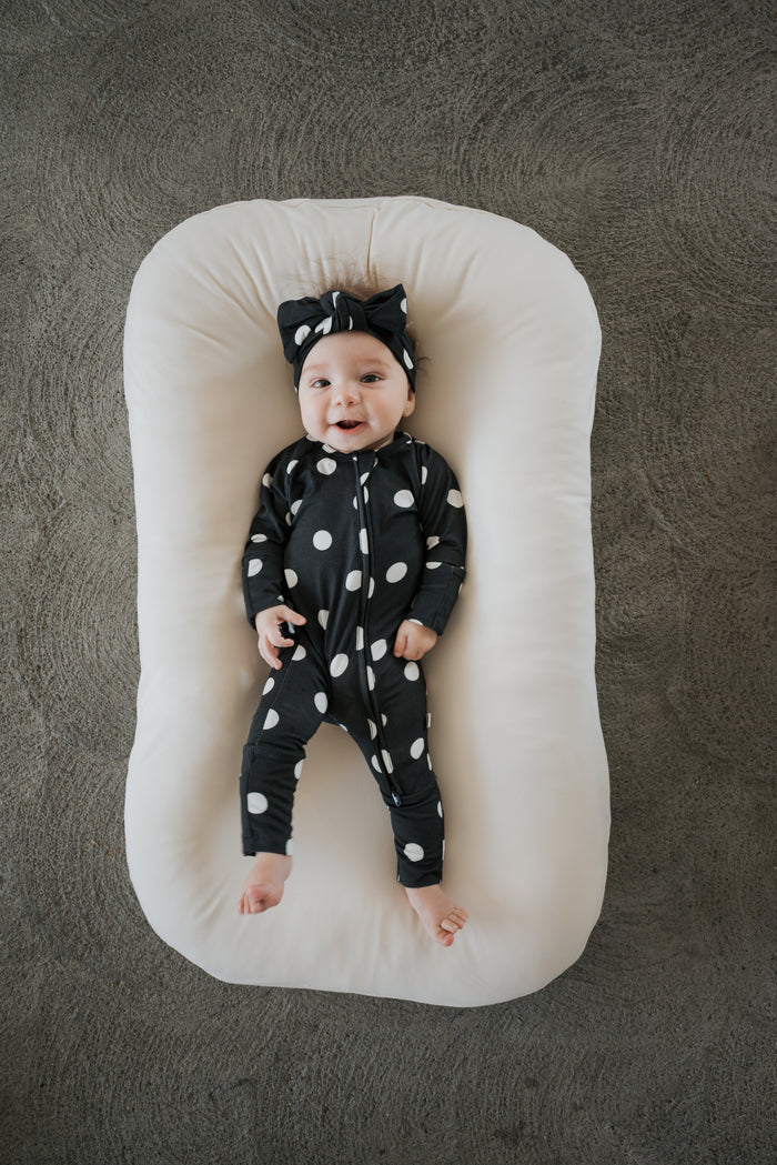 A smiling baby dressed in forever french baby’s Bamboo Zip Pajamas in B&W Polka Dot, complete with a matching headband, lies on a soft white cushion against a textured gray background.