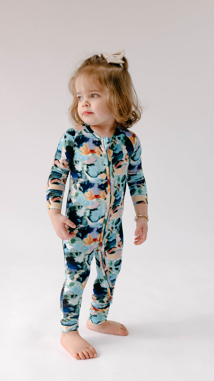 A toddler with light brown curly hair, wearing Forever French Baby's colorful Bamboo Zip Pajamas in Charli Print, stands barefoot against a plain white background. The child has a white bow in their hair, and the jumpsuit is made from soft bamboo fabric.