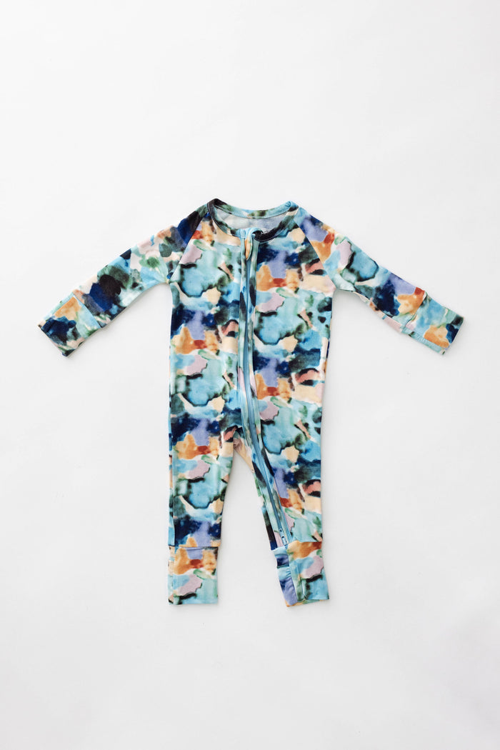 The Bamboo Zip Pajamas | Charli Print by forever french baby is a long-sleeved romper featuring a colorful abstract pattern with shades of blue, green, orange, and black. Made from soft bamboo fabric, this romper boasts a front zipper that extends from the neck to the left leg and has ribbed cuffs on both the sleeves and ankles. The garment is displayed laid flat on a white background.