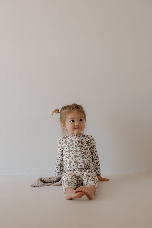 A young child with light hair in pigtails sits barefoot against a plain wall, wearing forever french baby Bamboo Zip Pajamas | Darlin', made from breathable bamboo fabric, and holding a small blanket.