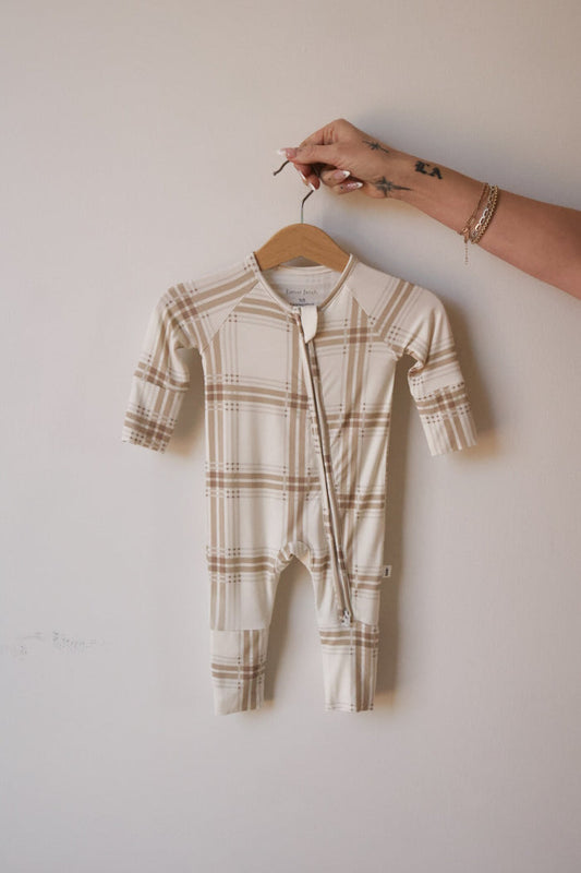 A hand holds forever french baby's Bamboo Zip Pajamas | Ellis Plaid, a beige and white plaid long-sleeve baby romper made from breathable fabric, against a plain light-colored wall.