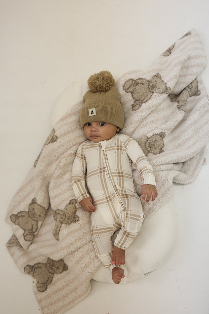 A baby in forever french baby’s Bamboo Zip Pajamas in Ellis Plaid and a tan pom-pom knit hat lies on a white cushion, partially covered by breathable bear-patterned sleepwear.