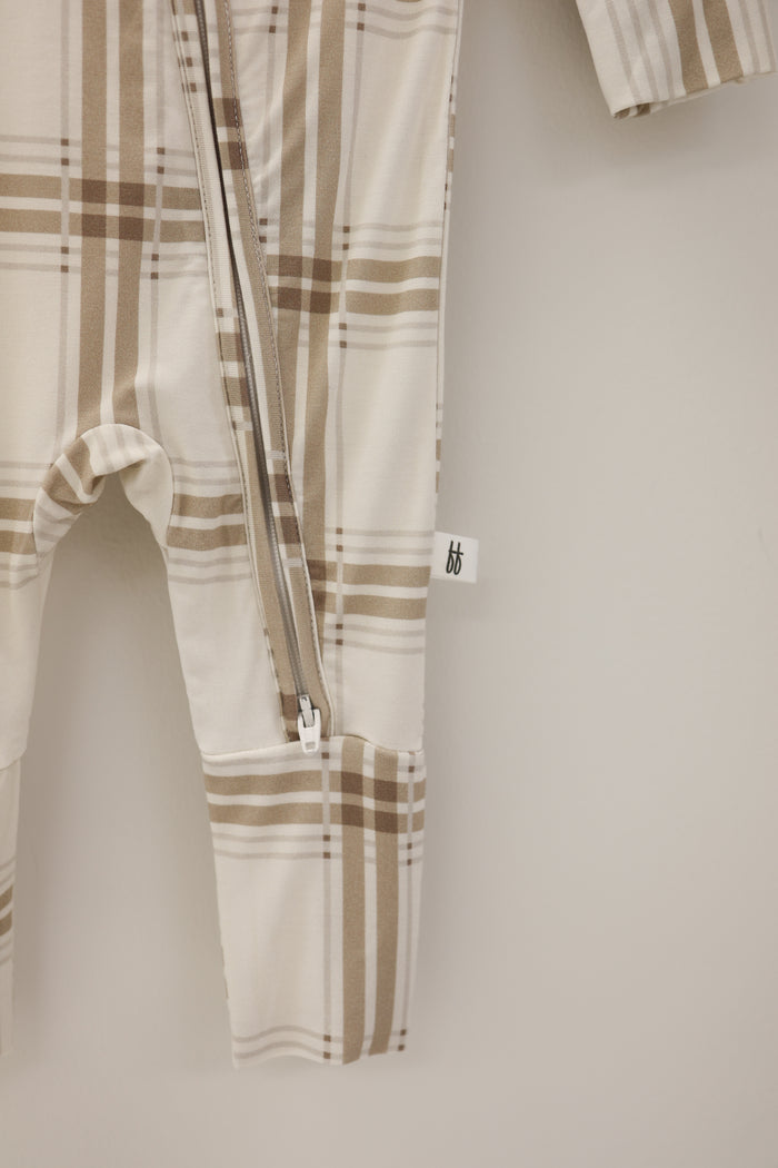 A close-up of the forever french baby Bamboo Zip Pajamas | Ellis Plaid shows a beige and brown plaid onesie with a diagonal zipper and side tag, made from hypoallergenic fabric, hanging against a light background.