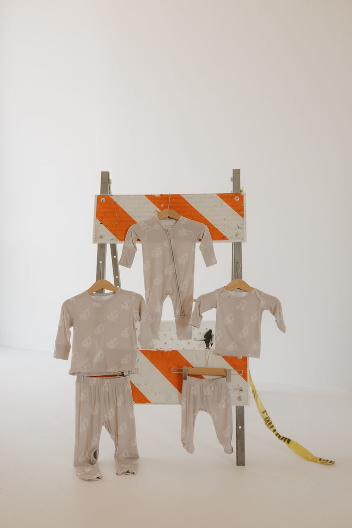 Four pieces of light beige Bamboo Zip Pajamas | Excavator by forever french baby, featuring white animal prints, hang on wooden hangers from an orange and white road barricade in a minimal white room.