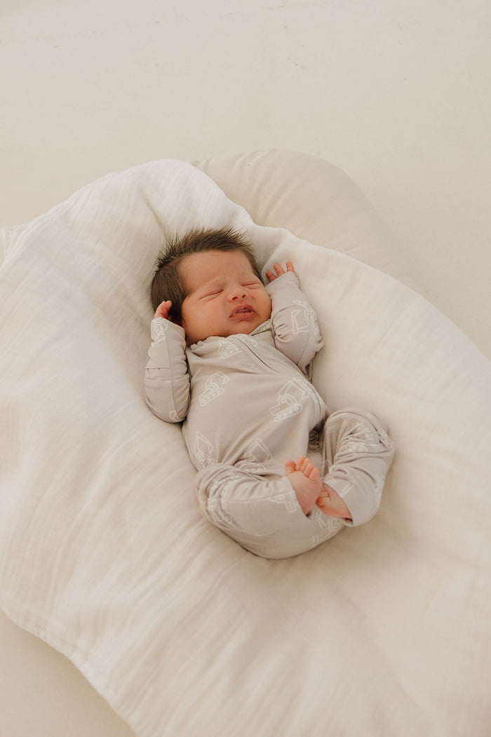 A baby wearing forever french baby's Bamboo Zip Pajamas | Excavator relaxes on a soft white blanket, arms up and eyes closed. The breathable bamboo material ensures comfort in this bright, tranquil setting.