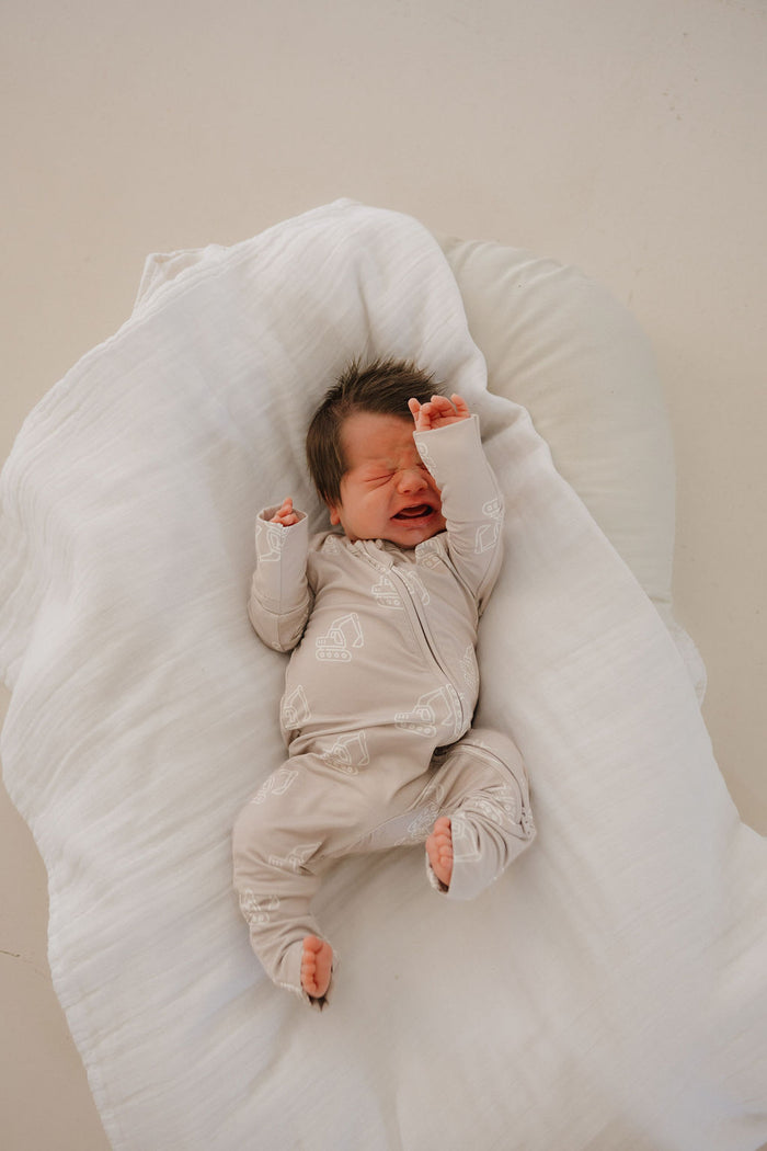 A newborn baby in forever french baby's Bamboo Zip Pajamas | Excavator lies on a white pillow, eyes closed with one arm raised, appearing to be crying or stretching.