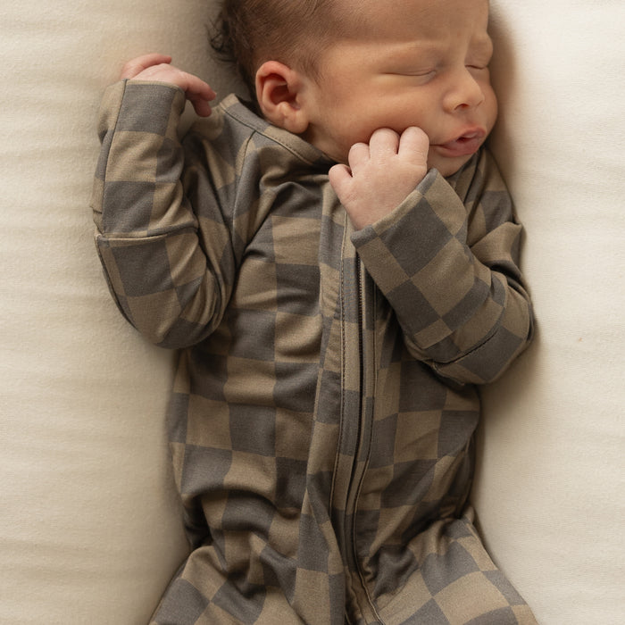 A newborn sleeps on a cream surface, wearing forever french baby's Bamboo Zip Pajamas in Faded Brown Checkerboard—a breathable, zippered onesie—resting peacefully with one hand by their face.