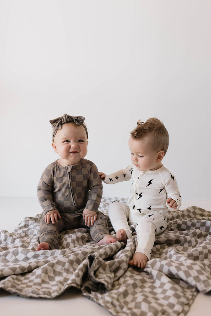 Two babies sit on a checkered blanket in a minimalistic space. The baby on the left, smiling with joy, wears the Bamboo Zip Pajamas in Faded Brown Checkerboard from forever french baby. The baby on the right, in a white hypo-allergenic onesie with black lightning bolts, reaches out towards the other baby.