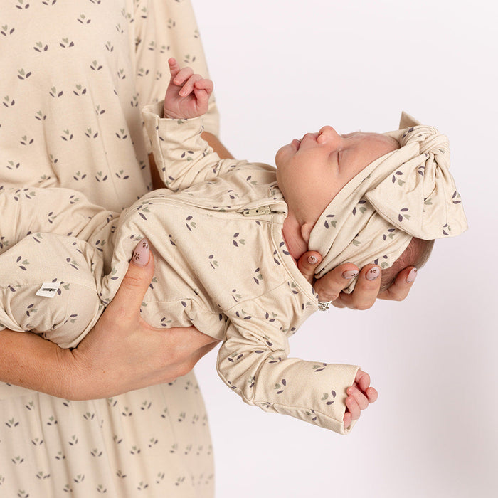 An adult holds a newborn dressed in forever french baby's Bamboo Zip Pajamas | Flora—a beige, leaf-patterned, breathable outfit with a matching headband—offering cozy comfort against a white background.