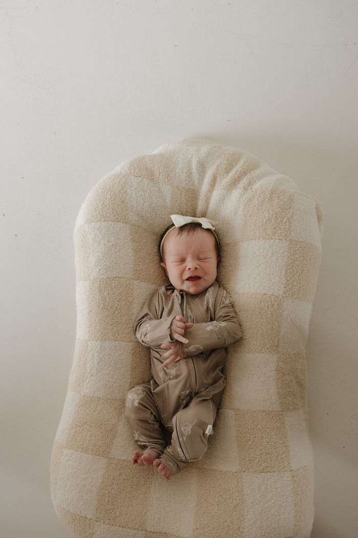 A newborn baby in forever french baby's Bamboo Zip Pajamas | Highland and a white bow headband lies on a soft, checkered beige and white cushion with eyes closed and hands clasped.