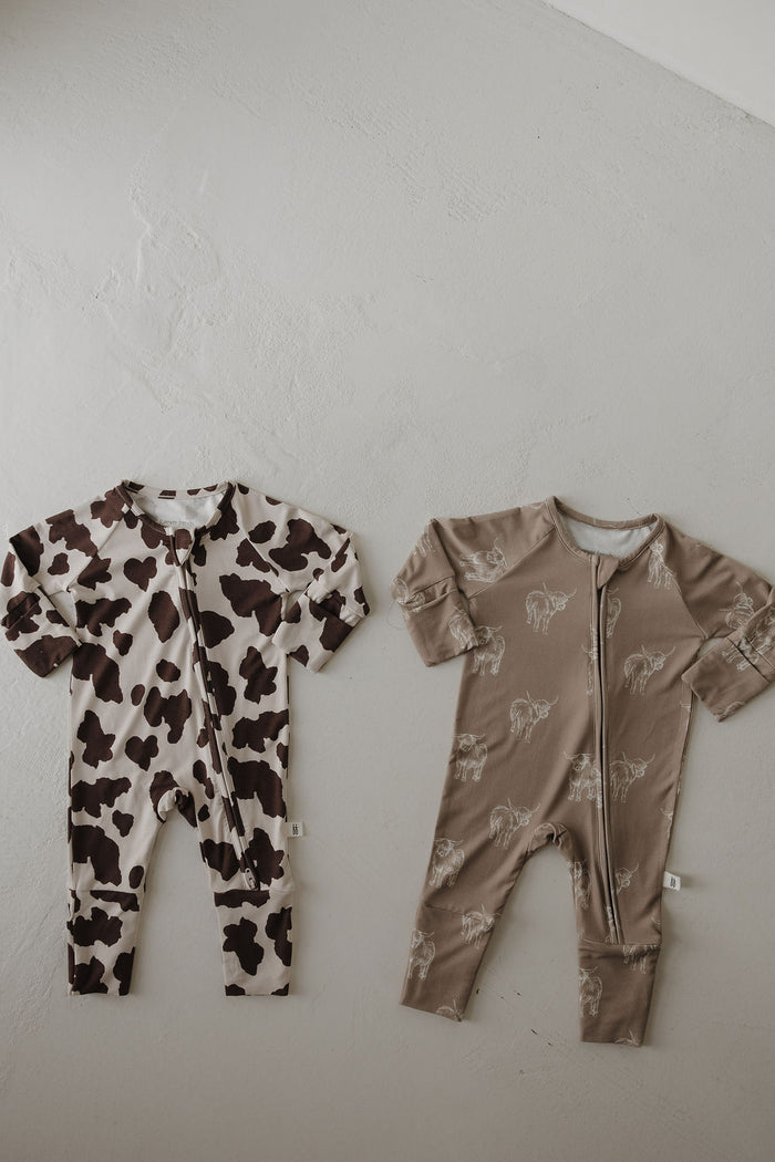 Two Bamboo Zip Pajamas by forever french baby are laid flat: one with a brown and white cow print, the other beige with small white animal outlines. These Highland pajamas offer breathable comfort for your baby all night long.