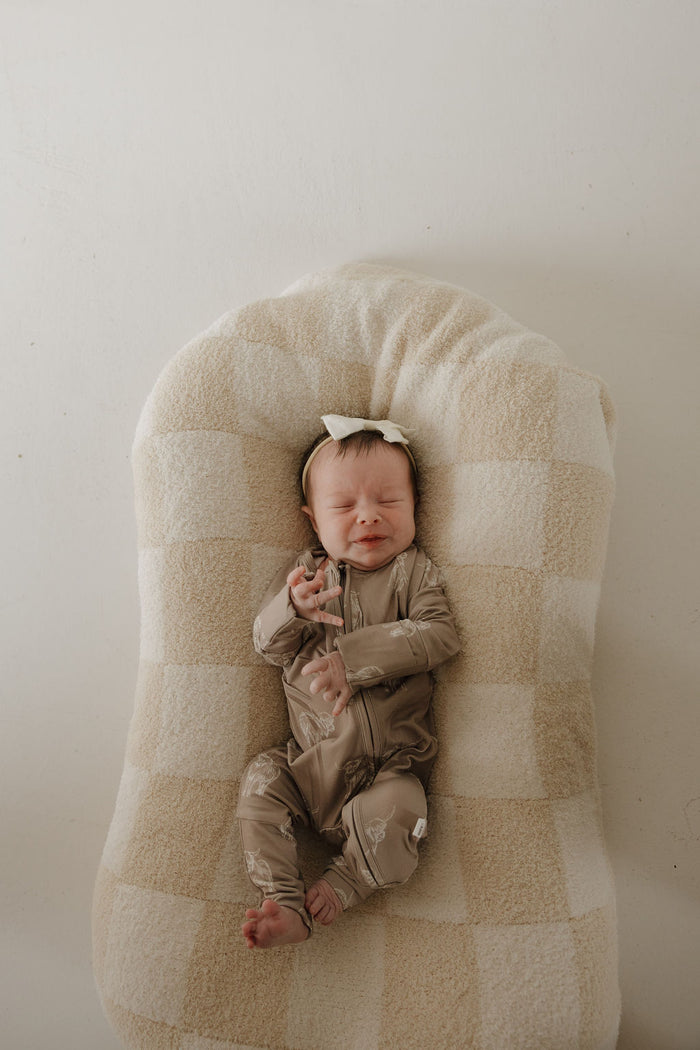A newborn in forever french baby’s Bamboo Zip Pajamas | Highland and a headband rests on a soft cream and tan checkered cushion, eyes closed, reaching upward.
