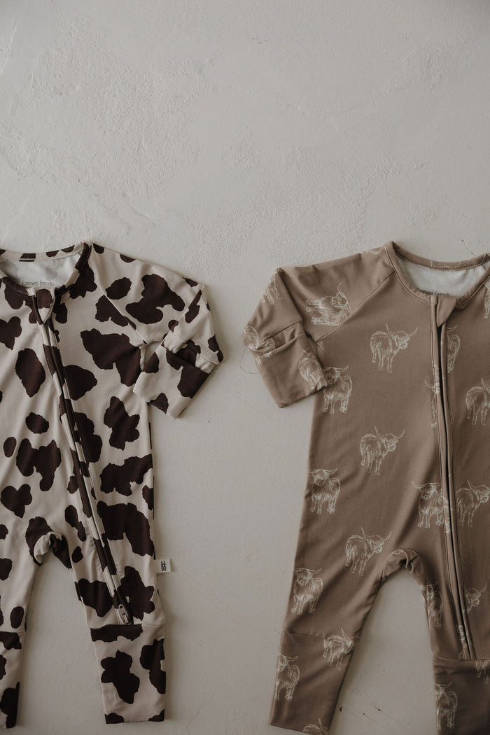 Two forever french baby Bamboo Zip Pajamas | Moo! on a light surface: one features a black and white cow print, the other is taupe with white line-drawn cows. Both have front zippers and long sleeves for easy comfort.
