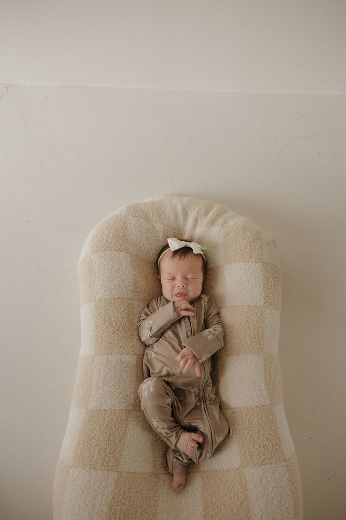 A newborn baby in forever french baby's Bamboo Zip Pajamas | Highland sleeps peacefully on a beige and white checkered cushion, wearing a white bow headband, against a plain background.
