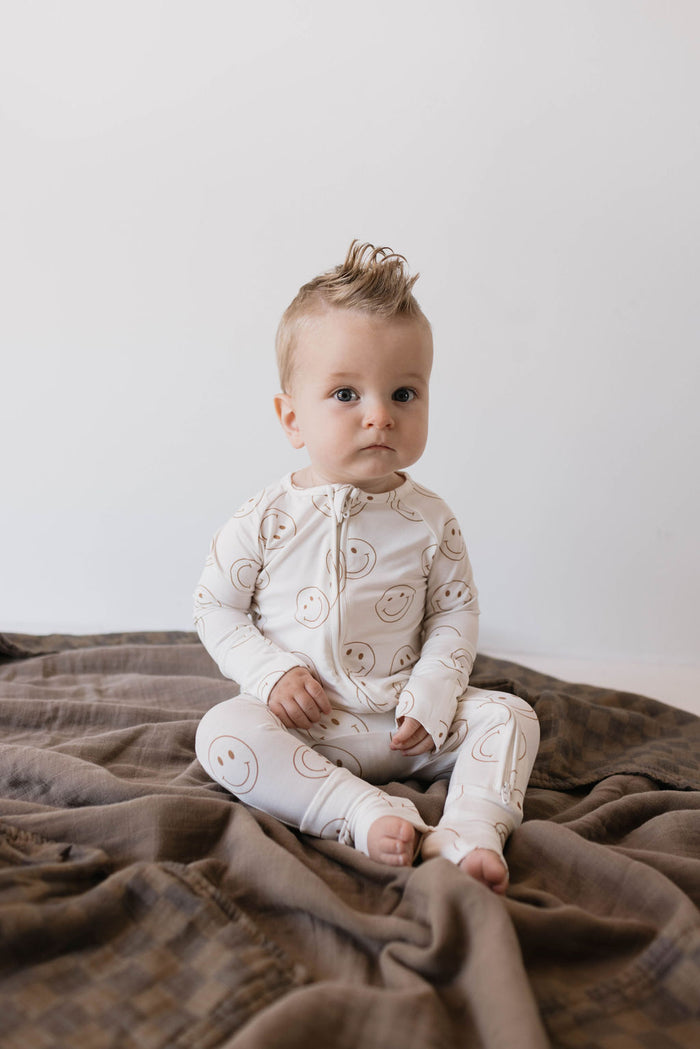 A baby with light-colored hair styled into a small mohawk is sitting on a brown blanket. They are wearing Forever French Baby's Bamboo Zip Pajamas in the 'Just Smile' pattern. The background is minimalistic with a white wall. The baby looks alert and focused.