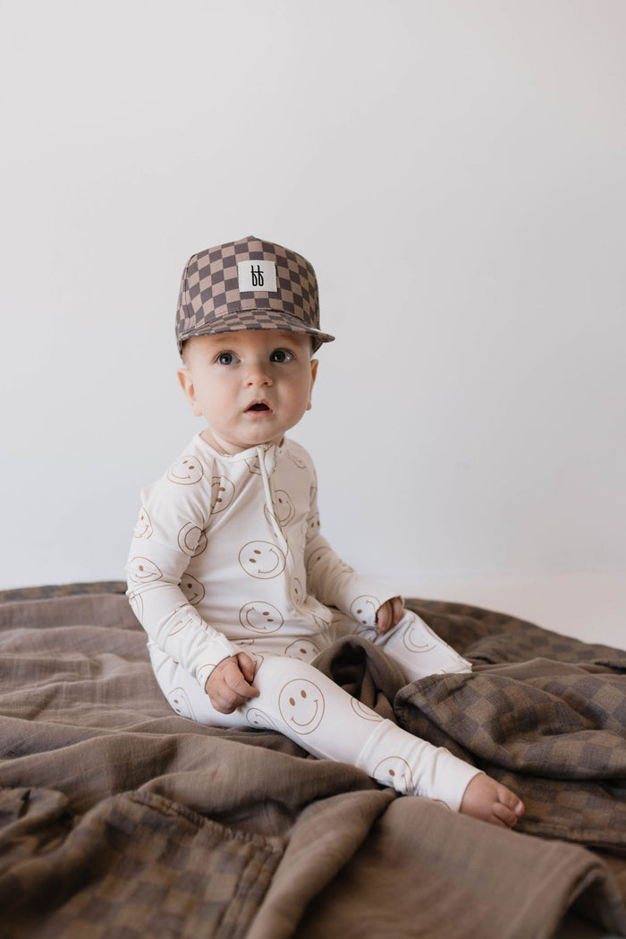 A baby is dressed in Forever French Baby's Bamboo Zip Pajamas in the "Just Smile" design, sitting on a brown blanket. The child is also wearing a checkered cap featuring a small logo on the front. The hypo-allergenic pajamas provide comfort for sensitive skin against the plain, light-colored background.