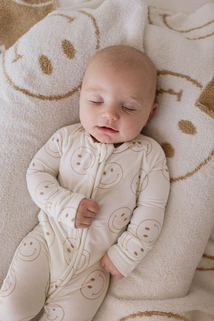 A baby dressed in Bamboo Zip Pajamas | Just Smile from forever french baby, adorned with brown smiley faces, is sleeping peacefully on a blanket that also features a smiley face pattern. The newborn clothing, crafted from premium bamboo fabric, ensures the ultimate comfort for the baby's calm and content sleep.