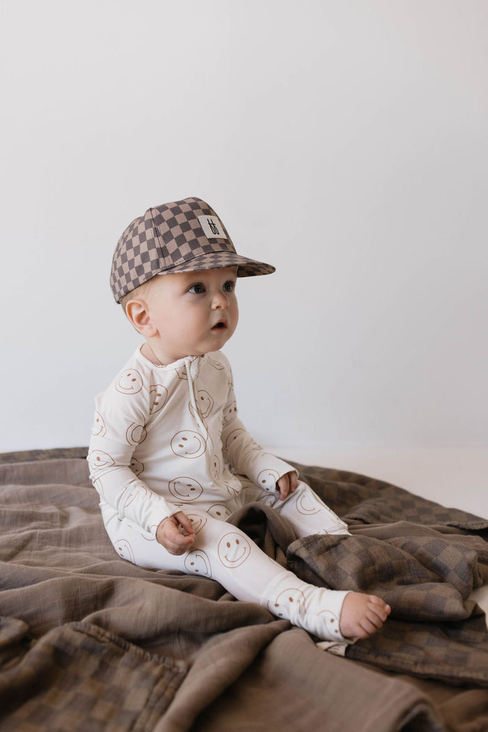 A baby in forever french baby's Bamboo Zip Pajamas | Just Smile, featuring a smiley face pattern, sits on a brown blanket. The baby wears a brown and beige checkered cap and looks to the side with an open-mouthed expression. The hypo-allergenic clothing ensures comfort, while the background is plain and light-colored.