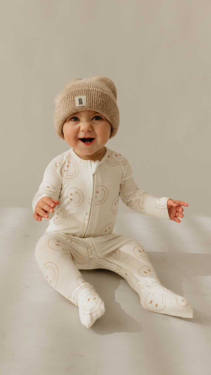 A baby wearing a beige knit hat and Bamboo Zip Pajamas | Just Smile by forever french baby, featuring smiley faces print, sits on a light-colored surface against a plain background. The baby is smiling, with one arm resting on the ground and the other reaching forward.