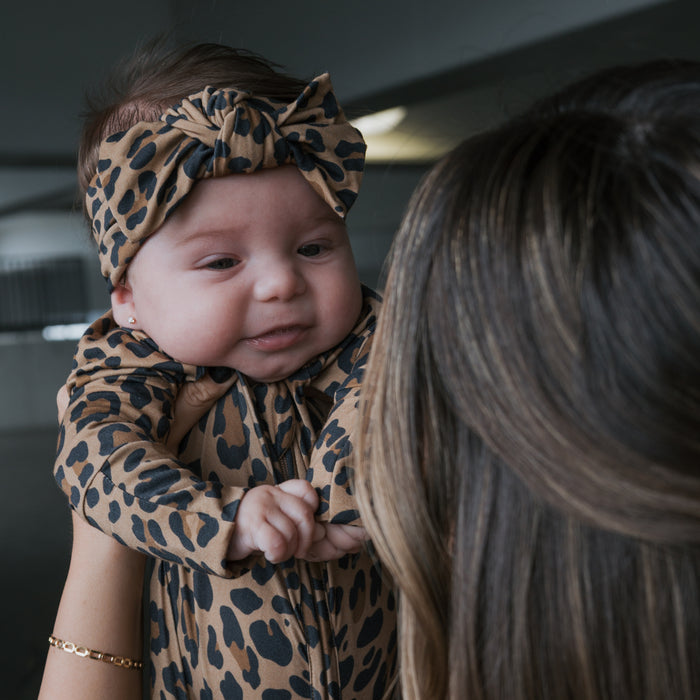 A baby wearing the forever french baby Bamboo Head Wrap | Leopard is held by an adult with long brown hair. The baby's gentle smile and the softly blurred background highlight the adorable head wrap.