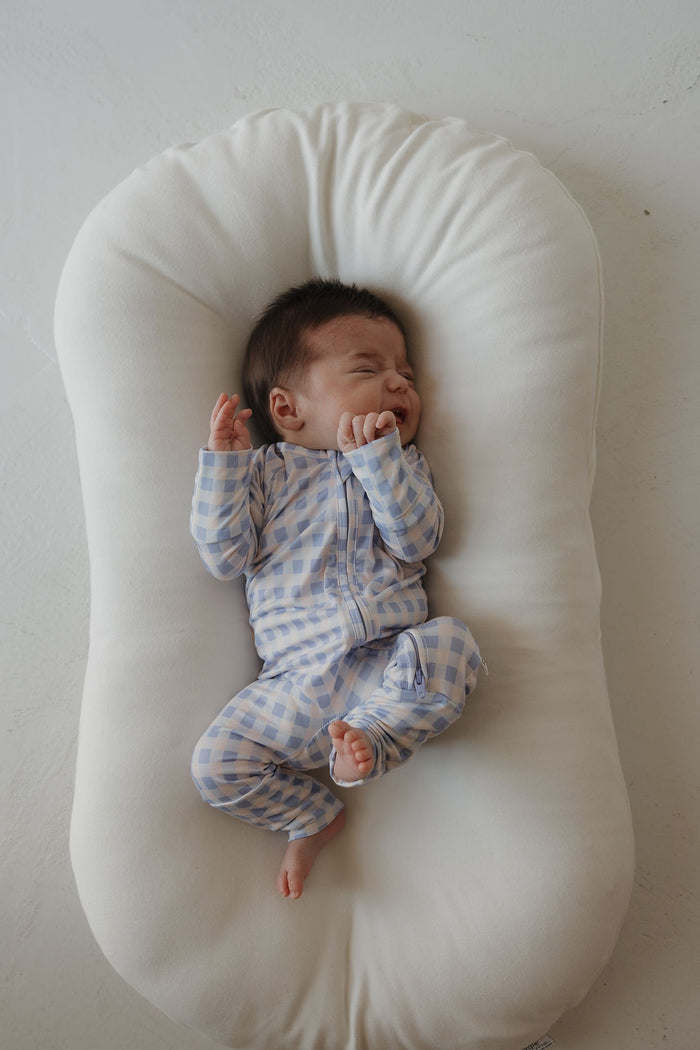 A baby dressed in the Marilyn Bamboo Zip Pajamas from forever french baby, featuring a blue and white checkered pattern, lies on a white cushioned pad with eyes closed and one hand near their face, appearing to cry or fuss. The hypoallergenic, breathable design ensures comfort for sensitive skin.