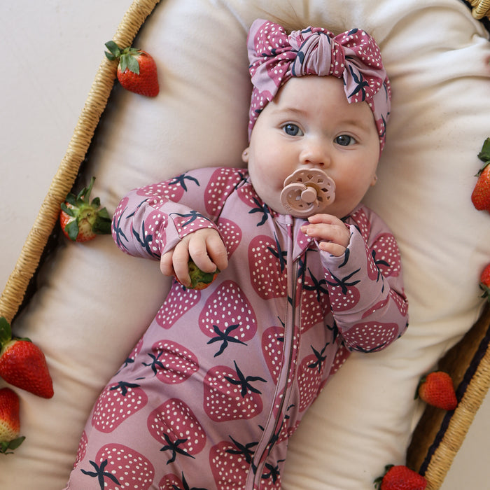 A baby in forever french baby's Bamboo Zip Pajamas | Matilda, wearing a pink strawberry-patterned headband, lies on a cushioned surface with fresh strawberries, holding one hand up and sucking on a pacifier.