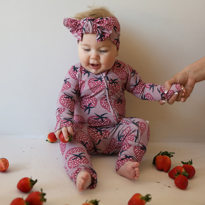 A baby in forever french baby’s Bamboo Zip Pajamas | Matilda—a pink, strawberry-patterned outfit with a matching headband—sits on a white surface, holding an adult’s hand, surrounded by fresh strawberries.