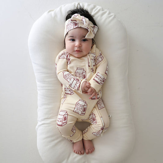 A baby rests on a white cushion, dressed in forever french baby's Bamboo Zip Pajamas | Milk—a cream sleepwear set with red milk carton prints and matching headband—hands folded, feet bare, gazing upward against a light backdrop.