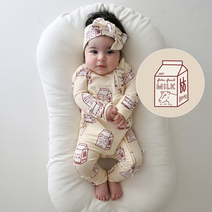 A baby lies on a white cushion, wearing forever french baby's Bamboo Zip Pajamas | Milk and a matching milk-carton headband. An illustrated milk carton sits beside the baby.