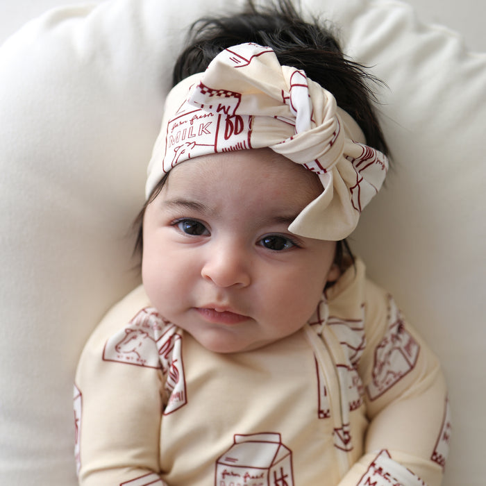 A dark-haired baby wears forever french baby Bamboo Zip Pajamas | Milk, featuring red milk carton prints, with a cream headband, resting on a white cushion and looking at the camera.