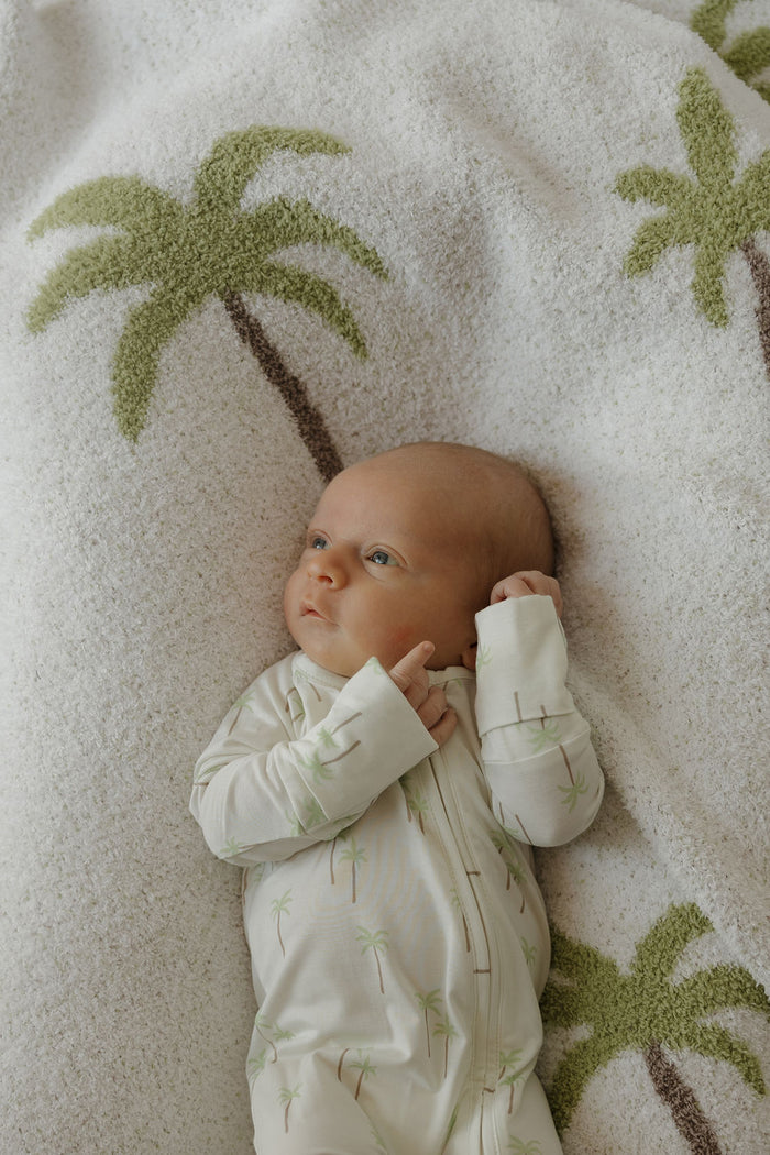 A baby in the forever french baby Bamboo Zip Pajamas | Palm, featuring a white fabric with green palm tree prints, lies on a soft white blanket adorned with green palms—comfortably dressed in breathable, gentle sleepwear ideal for delicate skin.