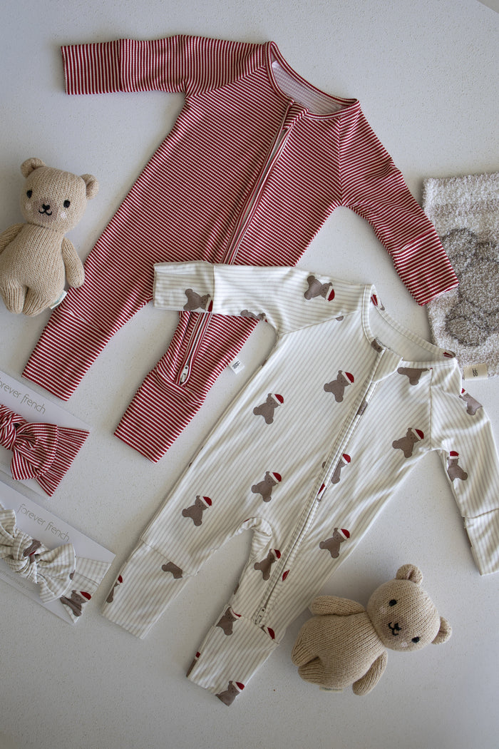 Two baby onesies from forever french baby are laid on a surface—one in the Peppermint Stripe Ribbed design and one white with a bear and hat pattern. Arranged around them are two small beige teddy bears, a bow headband, and a fuzzy washcloth. Crafted from ribbed bamboo fabric, these Bamboo Zip Pajamas are hypo-allergenic and provide both comfort and style for your little one.