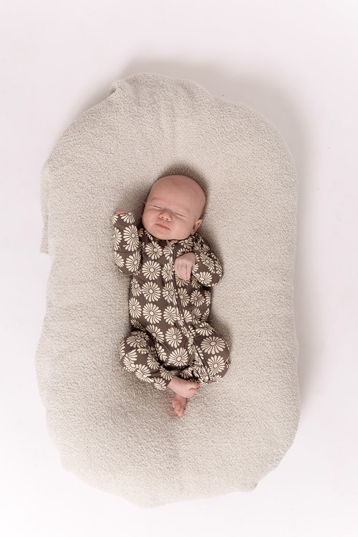 A baby sleeps in forever french baby Bamboo Zip Pajamas | Retro Floral—a brown, daisy-patterned onesie—on a soft light beige blanket, photographed from above on a white background.
