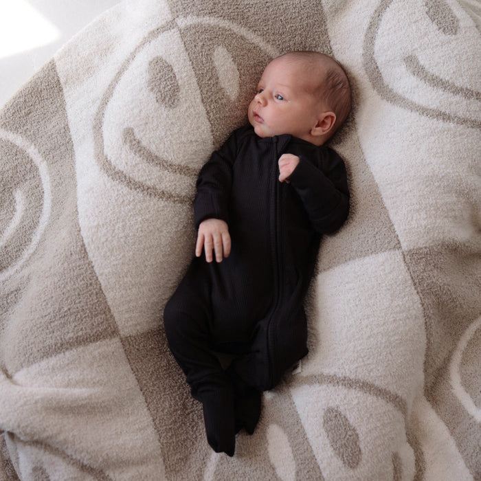 A newborn baby dressed in forever french baby's Bamboo Zip Pajamas in Black Ribbed lies on a cozy, hypo-allergenic blanket adorned with large smiley faces. The baby gazes to the side with a calm expression, enjoying the soft and light-colored background.